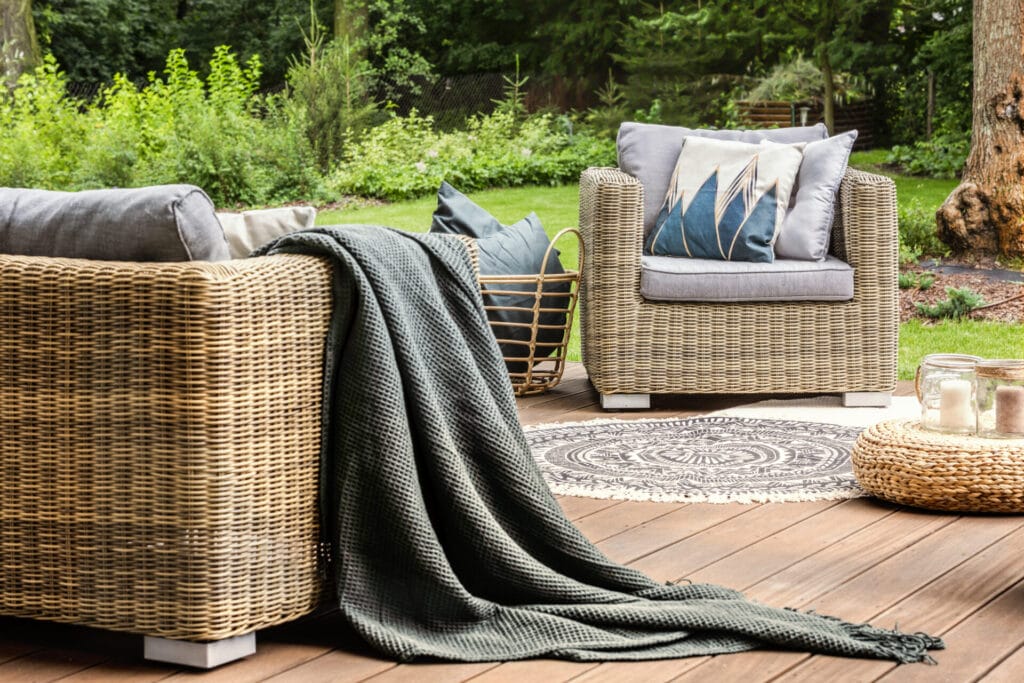 grey blanket on rattan sofa near armchair with pillows on wooden terrace with pouf. real photo